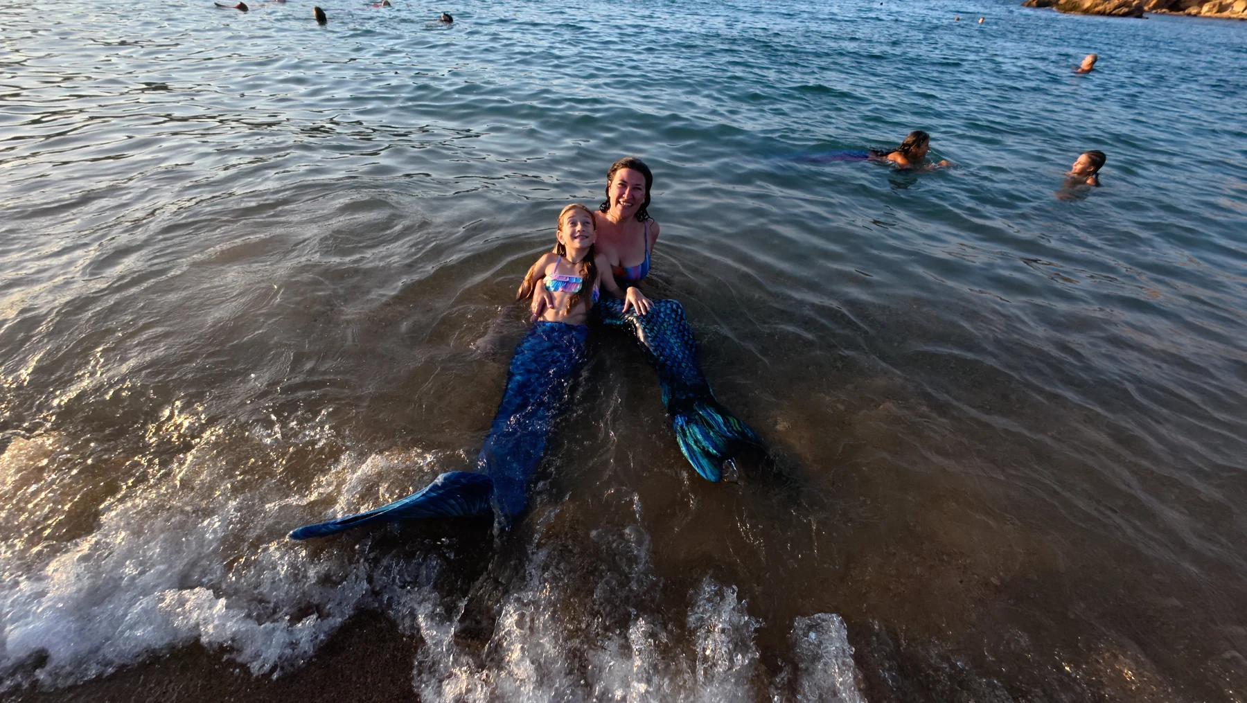 A mother and daughter smile together in blue mermaid tails