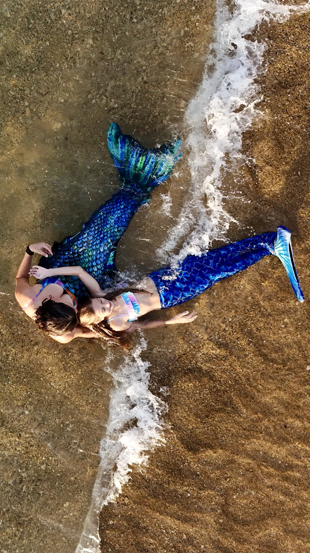 A mother cradles her daughter in mermaid tails beside a gentle wave